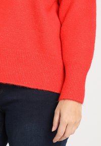 Rotem, strukturiertem Pullover mit gerippten Bündchen und Saum, entworfen für Komfort, kombiniert mit dunkelblauen Jeans im lässigen Schnitt.