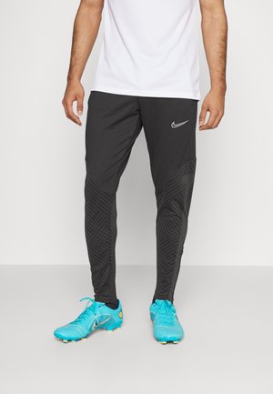 Mężczyzna w czarnych spodniach dresowych z logo Nike i jaskrawo niebieskich korkach piłkarskich stoi na szarej podłodze na białym tle.
