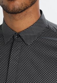 Czarna koszula zapinana na guziki z geometrycznym białym wzorem w kwadraty, z wyraźnie uformowanym kołnierzykiem i gładką teksturą materiału.