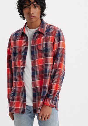 Camicia in flanella a quadri rossa e navy con due tasche sul petto, polsini con bottone e una vestibilità rilassata, indossata sopra una t-shirt bianca.