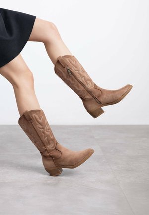 Botas western de ante marrón con un patrón decorativo cosido, cremallera lateral y tacón apilado. Con un diseño de punta redondeada.