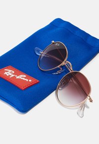 Sluneční brýle Ray-Ban v pilotním stylu s rámem z růžového zlata, gradientními tmavými skly a průhlednými nosními podložkami, leží na modré textilní tašce s červeným logem.