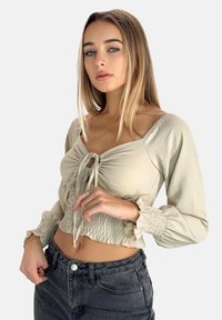 Ljusbeige kortärmad blus med knyt-fram detalj, långa puffärmar och smockad midja, ihop med högmidjade mörkgrå jeans.