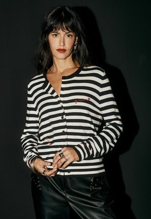 Donna con capelli neri e rossetto rosso indossa una camicia a righe bianco e nero abbottonata con dettagli rossi "love" e pantaloni di pelle neri.