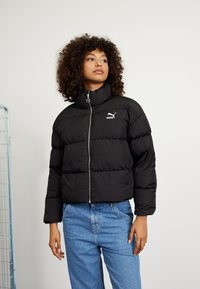 Svart puffjakke med høy krage, glidelås foran og logo. Boksformet med quiltet tekstur. Kombinert med blå denimjeans.