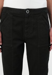 Pantalones de algodón negros con corte recto, que presentan bolsillos frontales en ángulo, un cierre de metal circular y detalles de costura visibles.