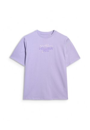 Młoda dziewczyna ubrana w lawendowy oversize T-shirt z napisem „Purrfect Fun”, szare szerokie spodnie, sportowe buty i kremową bluzę narzuconą na ramiona.