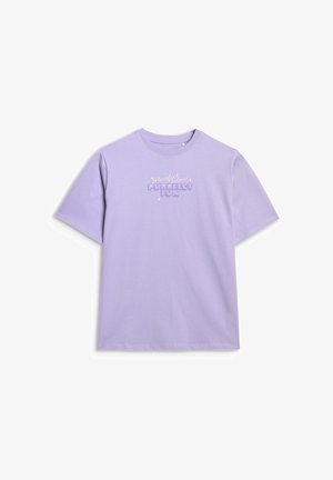 Młoda dziewczyna ubrana w lawendowy oversize T-shirt z napisem „Purrfect Fun”, szare szerokie spodnie, sportowe buty i kremową bluzę narzuconą na ramiona.