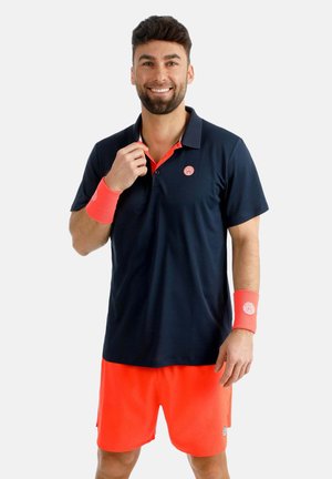 Uomo sorridente che indossa una polo blu navy, pantaloncini arancione acceso e braccialetti arancioni abbinati su entrambi i polsi.