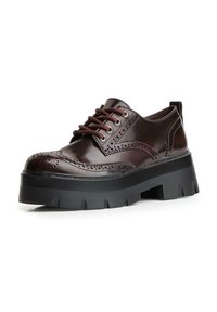 Scarpa brogue in pelle marrone scuro con perforazioni a wingtip, lacci marroni e suola spessa nera con tasselli su sfondo bianco.