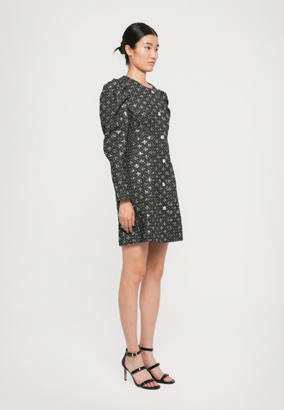 Woman standing wearing a black sequined long-sleeve mini dress with padded shoulders and black strappy high-heeled sandals.