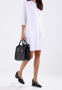 Robe chemise blanche à manches longues, sac à main noir texturé, et chaussures à talons noires. Lignes épurées et design minimaliste.