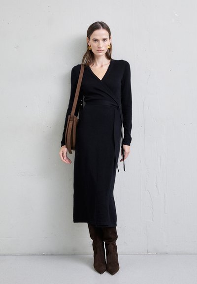 Robe noire en tissu tricoté avec des manches longues, dotée d'un décolleté en V profond et d'un lien à la taille. Associée à des bottes en daim marron au genou.