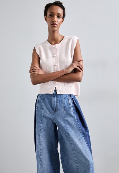Lichtroze mouwloze gebreide top met textuurstof en parelknopen, gecombineerd met wijde blauwe denim broek met contrasterende zijpanelen.