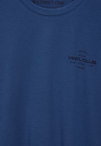 Sininen puuvillapaita, jossa on pyöreä kaula-aukko ja musta painettu logo, jossa lukee "VINYL CLUB" ja ylimääräistä tekstiä sekä koodeja ympyrämuodostelmassa.