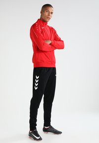 Rode sportjack met ritssluiting aan de voorkant, zwarte trainingsbroek met een wit chevronpatroon, en zwarte sportschoenen met rode accenten.