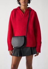 Maglione rosso lavorato a maglia con scollo a V e polsini a costine, abbinato a una borsa a tracolla in pelle nera con patta curva e dettaglio del logo.