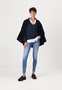 Marineblå genser over en hvit skjorte med krage, kombinert med lyseblå skinny jeans og svarte loafers. Modellen har på seg en mørk jakke og øreringer.