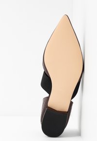 Chaussure brune à bout pointu avec un dessus en cuir texturé, une sangle élastique noire et une semelle de couleur claire, légèrement texturée.