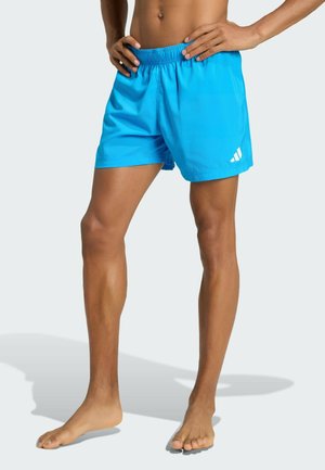 Mandligt torso og ben iført lyseblå sportsshorts med lille hvidt logo på venstre ben, står barfodet med hænderne på hofterne.