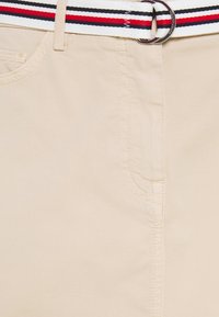 Tela beige con una textura suave, que presenta un bolsillo delantero y un cinturón a rayas en rojo, negro y blanco con una hebilla metálica.