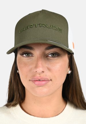 Femme aux cheveux châtain clair et aux yeux verts portant une casquette vert olive et blanche avec « GERTRUDE » brodé à l'avant.