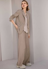 Beige op maat gemaakt blazer van lichte stof, gecombineerd met een bijpassende blouse en wijdvallende broeken. Chunky witte sneakers maken de outfit compleet.