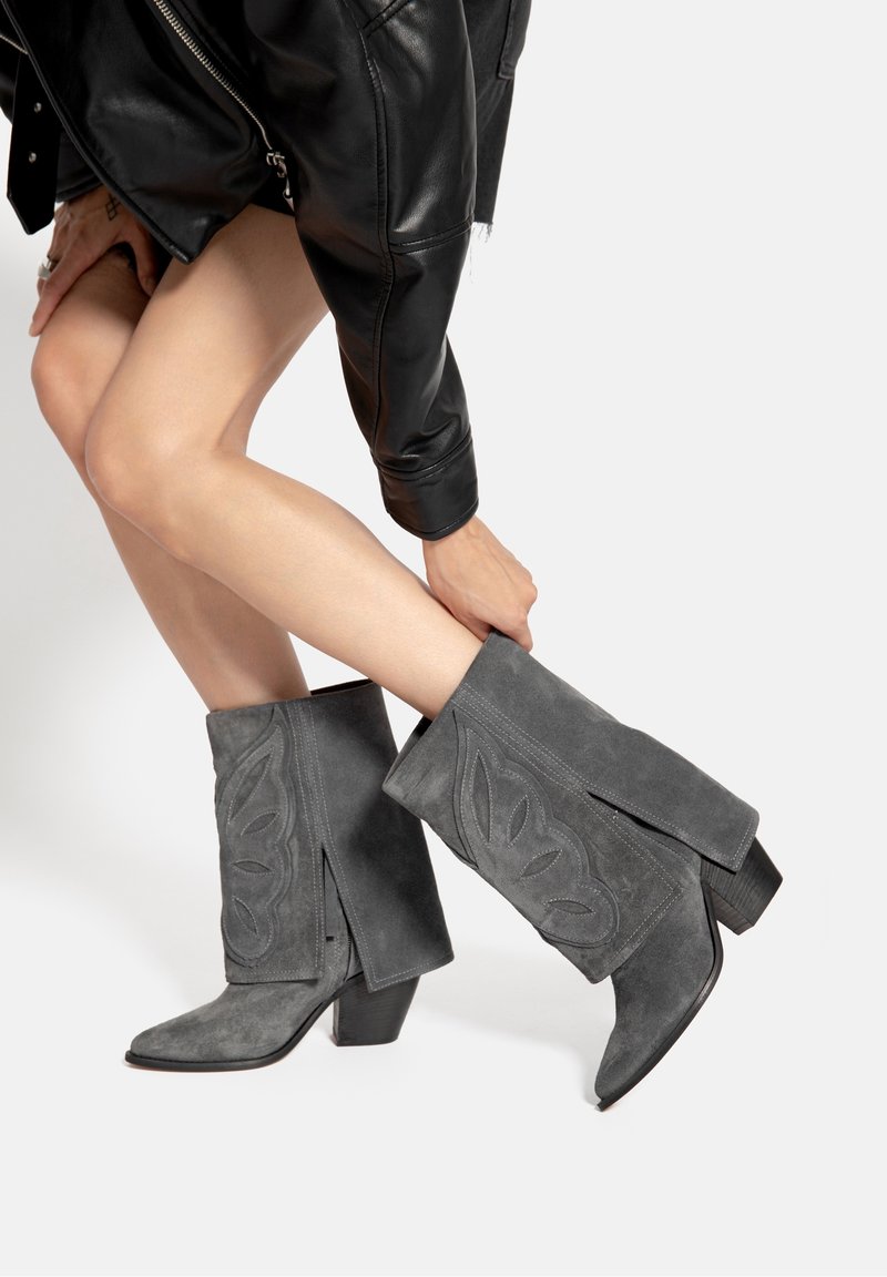 Personne ajustant des bottes de cowboy en daim gris avec broderies de feuilles, portant une veste en cuir noire et une jupe courte sur fond blanc.