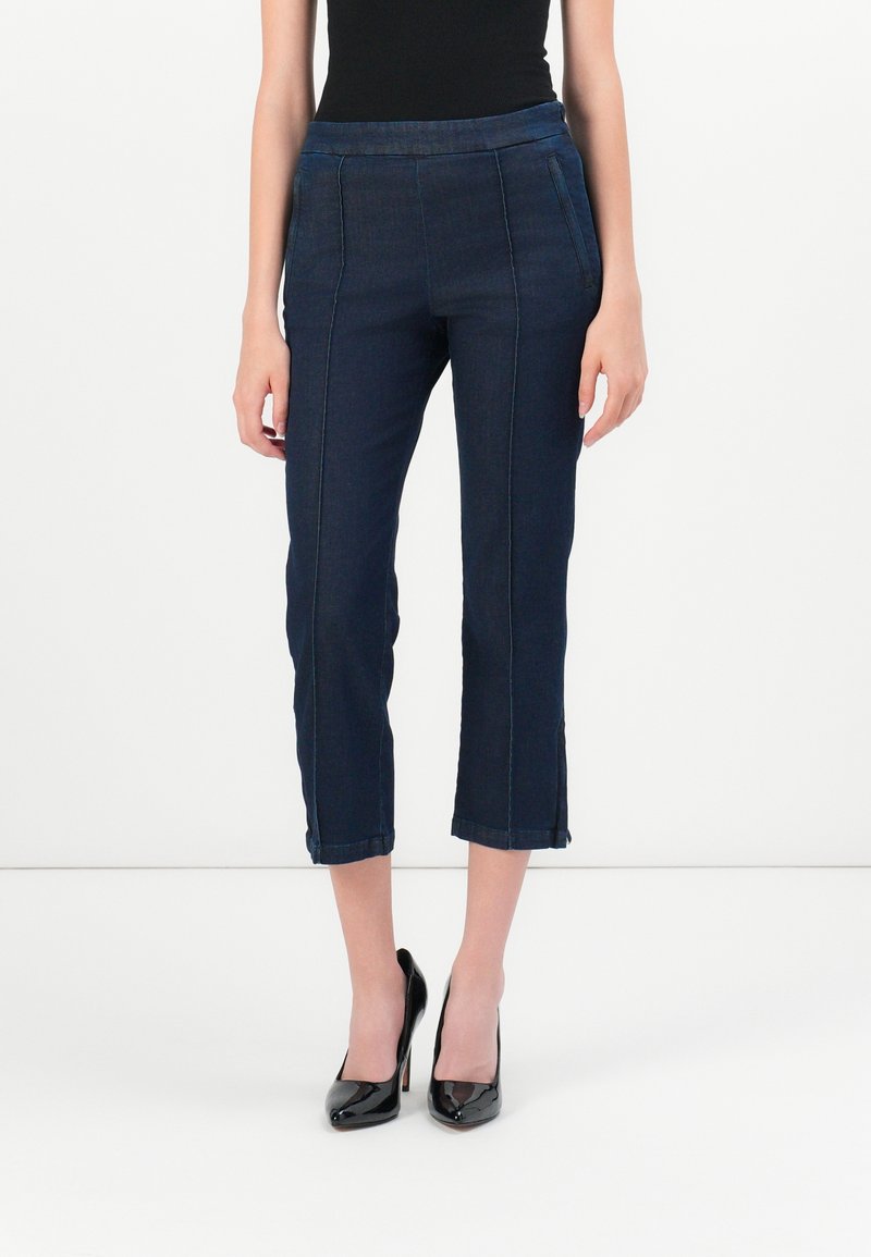 Pantalon court en denim bleu foncé avec taille ajustée, devant plat, poches latérales et fentes aux ourlets, associé à des chaussures à talons hauts noires.