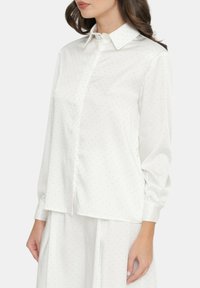 Camicia bianca con bottoni e maniche lunghe, caratterizzata da un sottile motivo a pois e un colletto classico. Tessuto morbido e vestibilità comoda.