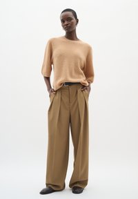 Beige gestrickter Pullover mit kurzen Ärmeln, kombiniert mit locker sitzenden, hochtaillierten braunen Hosen mit Falten, ergänzt durch einen schwarzen Gürtel.