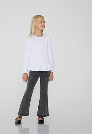 Langärmliges weißes Shirt mit Rüschensaum, kombiniert mit ausgestellten schwarzen Jeans. Schwarze Ballerinas vervollständigen das Outfit. Einfaches und modernes Design.