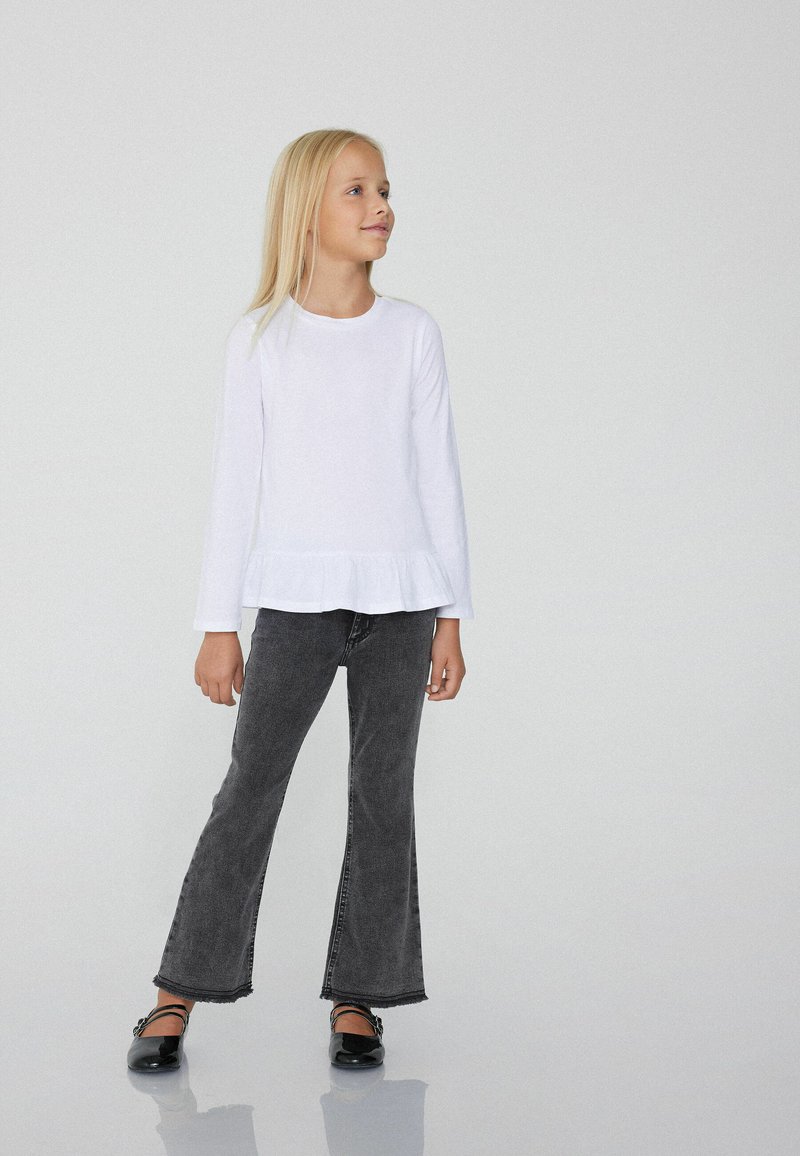 Bílá košile s dlouhými rukávy a volánkovým lemem, spárovaná s rozšířenými černými džíny. Černé baleríny doplňují outfit. Jednoduchý a moderní design.