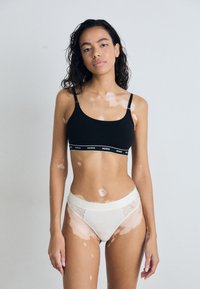 Bralette noire avec fines bretelles, dotée d'une bande contrastante portant l'inscription "HUGO." Culotte de bikini blanche avec des panneaux en tulle à pois et une ceinture douce.