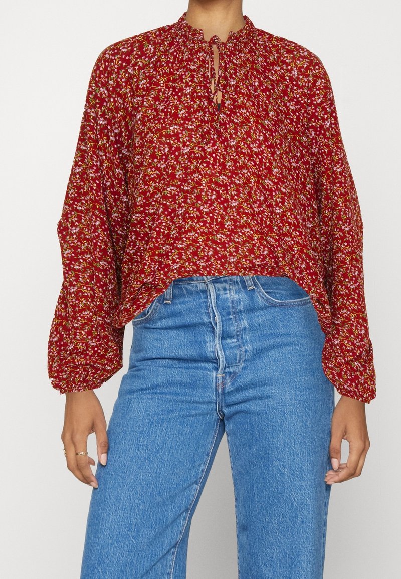 Personne portant un chemisier rouge ample à motifs floraux avec des manches longues et un jean bleu taille haute, debout devant un fond blanc.