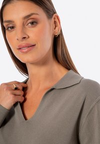 Beige Pulli mit V-Ausschnitt und weicher Textur, mit einem Kragen versehen, getragen mit einem goldenen Ring an einer Hand, die auf der Schulter ruht, mit dezenten Details.