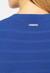 Modrý pletený top s horizontální texturou v rib strukturu. Na zadní výstřihu je viditelný kovový štítek s názvem značky pro detail.