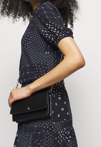 Černá kožená crossbody kabelka se zlatým logem, s klopou, řetízkem a hladkou texturou. Nosit s tmavými vzorovanými šaty.