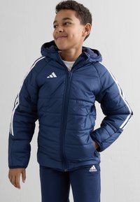 Granatowa kurtka puchowa z białymi paskami na rękawach i logo Adidas, z kapturem, zamkiem błyskawicznym i pikowaną teksturą.