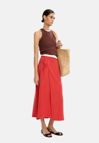 Brązowy top bez rękawów, biały podkoszulek, czerwona spódnica z wiązaniem, czarne sandały i pleciona torba ze słomy. Prosty, czysty design.