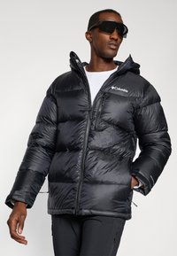 Schwarze Pufferjacke mit Reißverschluss, Kapuze und Columbia-Logo. Glattes, glänzendes Material mit gestepptem Design.