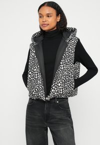 Γιλέκο με επένδυση και μοτίβο ασπρόμαυρων κηλίδων, κλείσιμο με φερμουάρ και επενδυμένο κουκούλι. Φορεμένο πάνω από ένα μαύρο ζιβάγκο και σε συνδυασμό με σκούρο τζιν.