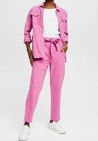 Giacca di jeans rosa con chiusura a bottoni e due tasche sul petto, abbinata a pantaloni rosa con cintura e sneaker bianche.