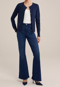 Cardigan blu navy con bottoni dorati, abbinato a jeans flare in denim scuro a vita alta e scarpe con tacco nere. Canotta bianca a costine sotto.