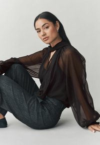 Čierna priesvitná blúzka s detailom šatky, dlhými puffovými rukávmi a výstrihom do V. Tmavosivé textúrované nohavice, pózovanie v sede na svetlom pozadí.