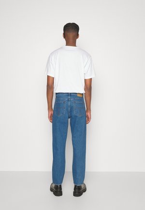 Person steht mit dem Rücken zur Kamera, trägt ein einfarbiges weißes T-Shirt, blaue Jeans und schwarze Schuhe vor einem einfarbigen weißen Hintergrund.