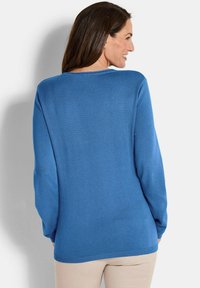 Eine Frau lächelt, schaut weg, trägt einen blauen Pullover mit langen Ärmeln und beigen Hosen vor einem schlichten weißen Hintergrund.