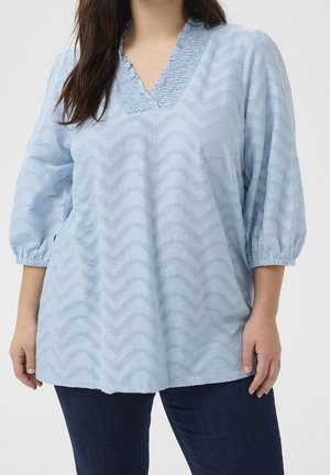 Kobieta w jasnoniebieskiej teksturowanej bluzce z dekoltem w serek i rękawami do połowy przedramienia oraz ciemnoniebieskich dżinsach, stojąca na jednolitym tle.