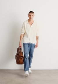 Kremfarget ribbestrikket genser, blå jeans og hvite sneakers, bærende en brun lærduffelbag, gående på et grått gulv mot en hvit vegg.