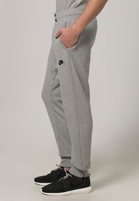 Grå Nike sweatpants i mjuk fleece med taperad passform, sidofickor, dragsko i midjan och svart logo på vänster lår. Svarta sneakers synliga.